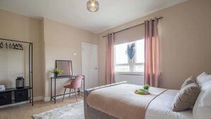 A bedroom with a bed, a desk, a chair, a window with curtains, and a hanging light fixture. - Mitchell Apartment - 1 Bedroom (North Lanarkshire)