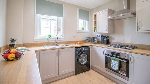 A modern kitchen with a dishwasher, oven, microwave, and a fruit bowl on the counter. - Mitchell Apartment - 1 Bedroom (North Lanarkshire)