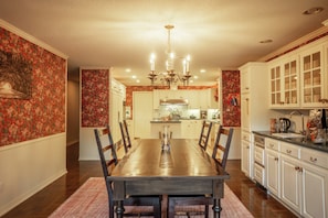 A dining area with a wooden table and chairs, a chandelier, and a kitchen in the background.