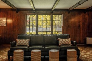 A room with a dark leather sofa, three woven ottomans, a large window with a view of trees, and wooden walls.