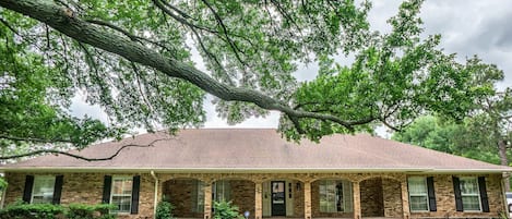 A two-story brick house with a front porch, a garage, and a large tree in front.