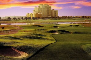 Ein Golfplatz mit Bunkern und einem Wasserschutzhindernis, im Hintergrund ein Hotel und ein Sonnenuntergangshimmel.