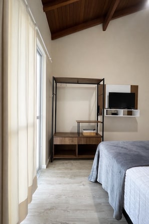Ein Schlafzimmer mit einem Bett, einem an der Wand befestigten Fernseher, einem Holztisch und einem Fenster mit Vorhängen.