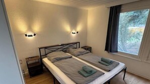 A double bed with two pillows, a nightstand, a window with curtains, and wall-mounted lighting. - Vacation village Seddiner See - Bungalow 6 (Seddiner See)