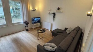 A living room with a sofa, a TV, a small table, and a window with curtains. - Vacation village Seddiner See - Bungalow 6 (Seddiner See)