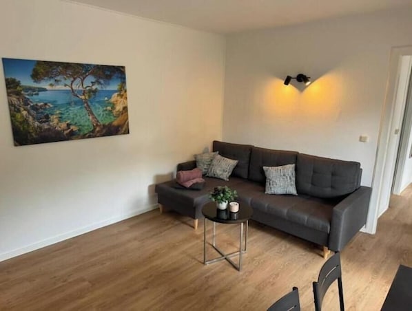 A living room with a grey sofa, a small round table with a plant, and a painting of a coastal scene.