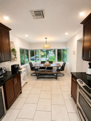 A modern kitchen with a dining table, dark wooden cabinets, a built-in oven, and a large window offering a view of greenery. - Miami World Cup Pool House Near Hard Rock Stadium • 5 Min to Beach (Hollywood)