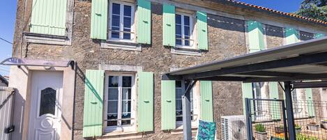 Un bâtiment à deux étages avec des volets verts, une porte blanche et un patio couvert agrémenté de chaises et de plantes en pot.