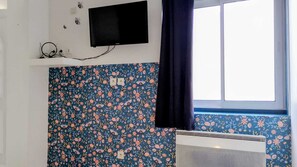 Ein Zimmer mit blumenmusterbehangener Wand, einem angebrachten Fernseher und einem Fenster mit Vorhang.