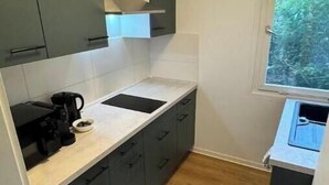 Une cuisine compacte avec des meubles gris, un plan de travail blanc, une machine à café et une fenêtre donnant sur un espace verdoyant.