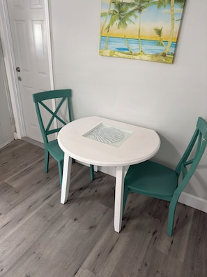 Ein runder, weißer Tisch mit einem Ventilator, zwei türkisfarbenen Stühlen und einem Gemälde mit Strandmotiv an der Wand.