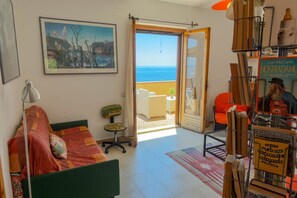 A room with a bed, a chair, a desk, a bookshelf, a painting, and a view of the sea. - Casa Ilune (Cala Gonone)