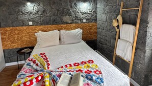 Een slaapkamer met een houten plafond, stenen muren, een bed met een kleurrijke deken, een trap met handdoeken en een klein tafeltje met een boek.