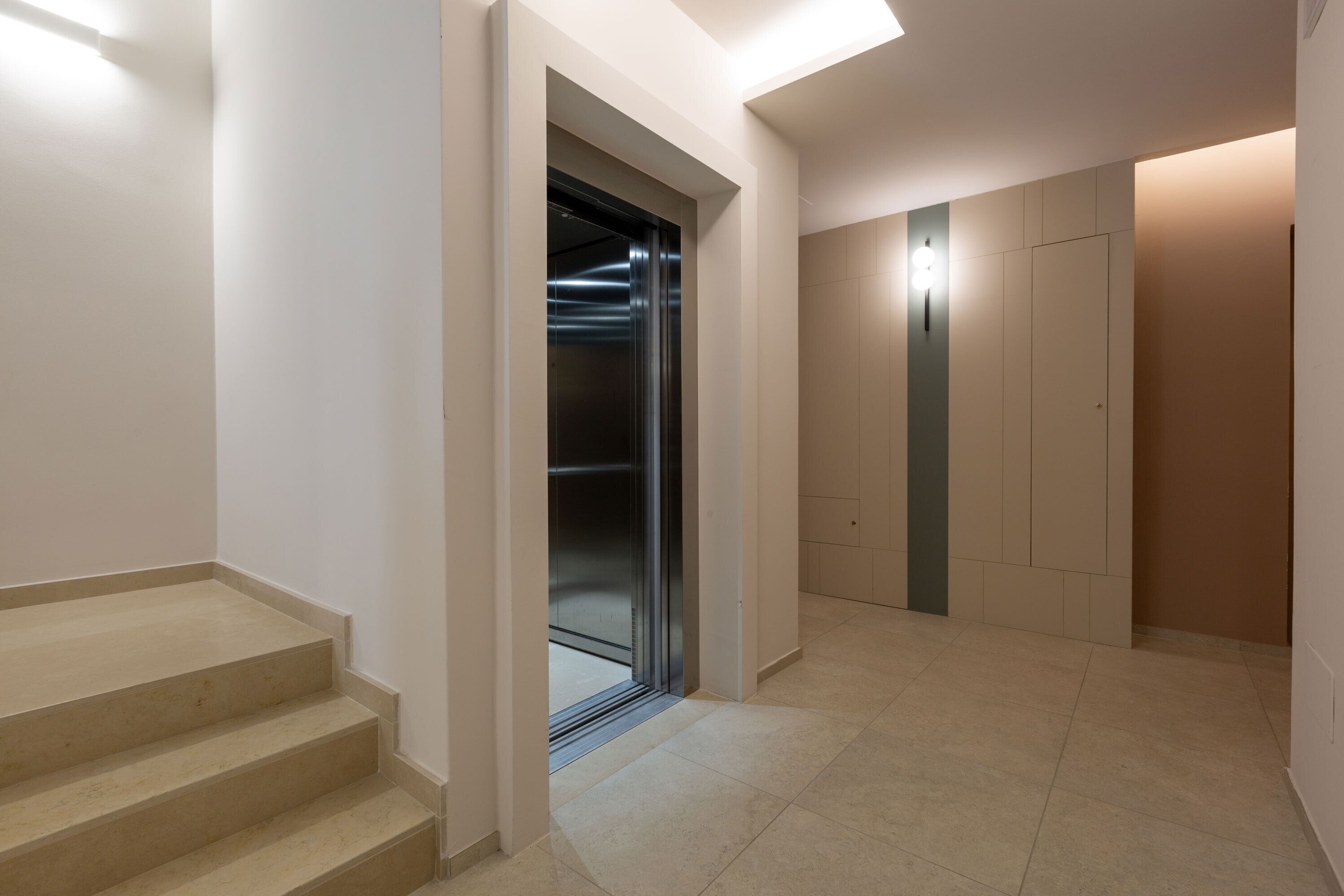 A hallway with a staircase, a door with a metallic handle, and a light fixture on the wall.