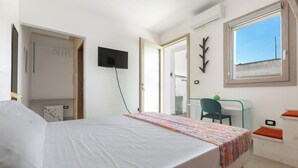 A bedroom with a bed, a desk, a chair, a TV, and a window.