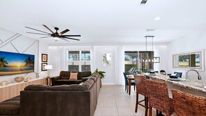Un salon moderne avec un canapé, un coin repas avec des chaises en osier, une cuisine avec un bar et un ventilateur de plafond.