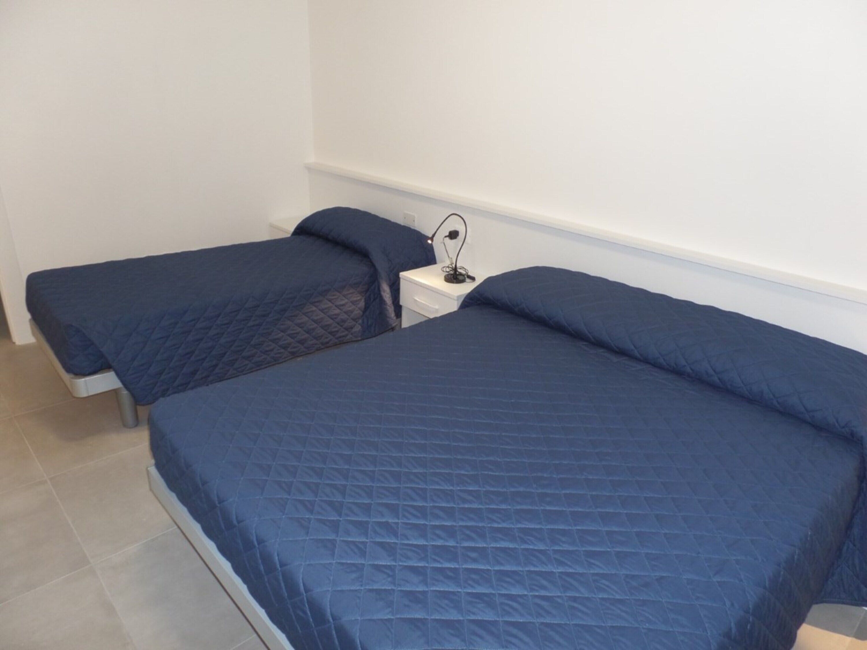 Quarto com duas camas, cada uma com colcha azul acolchoada, mesa de cabeceira branca com abajur e parede branca lisa.