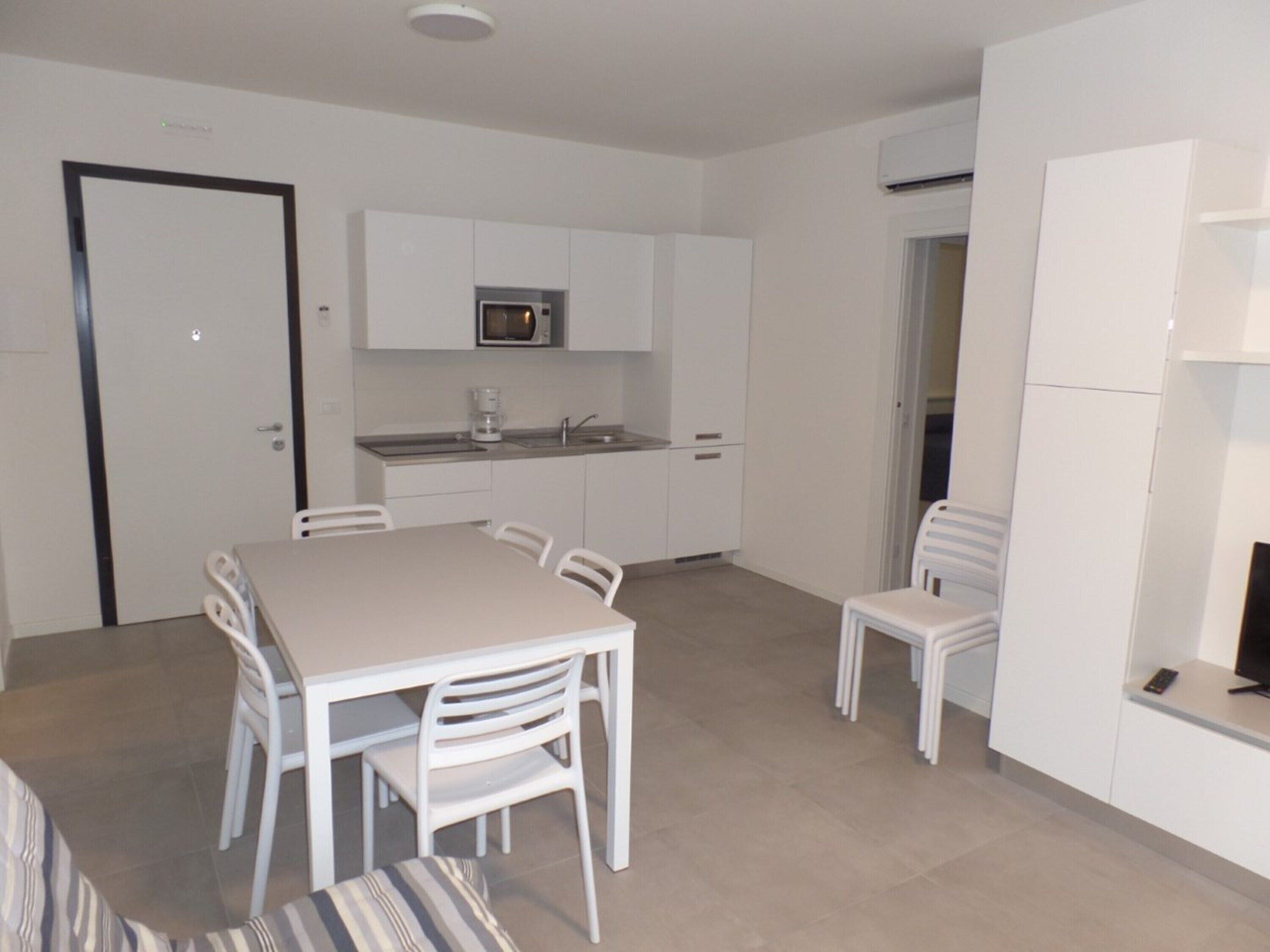 A modern kitchen with white cabinetry, a microwave, and a coffee maker. There is a dining area with a table and chairs.