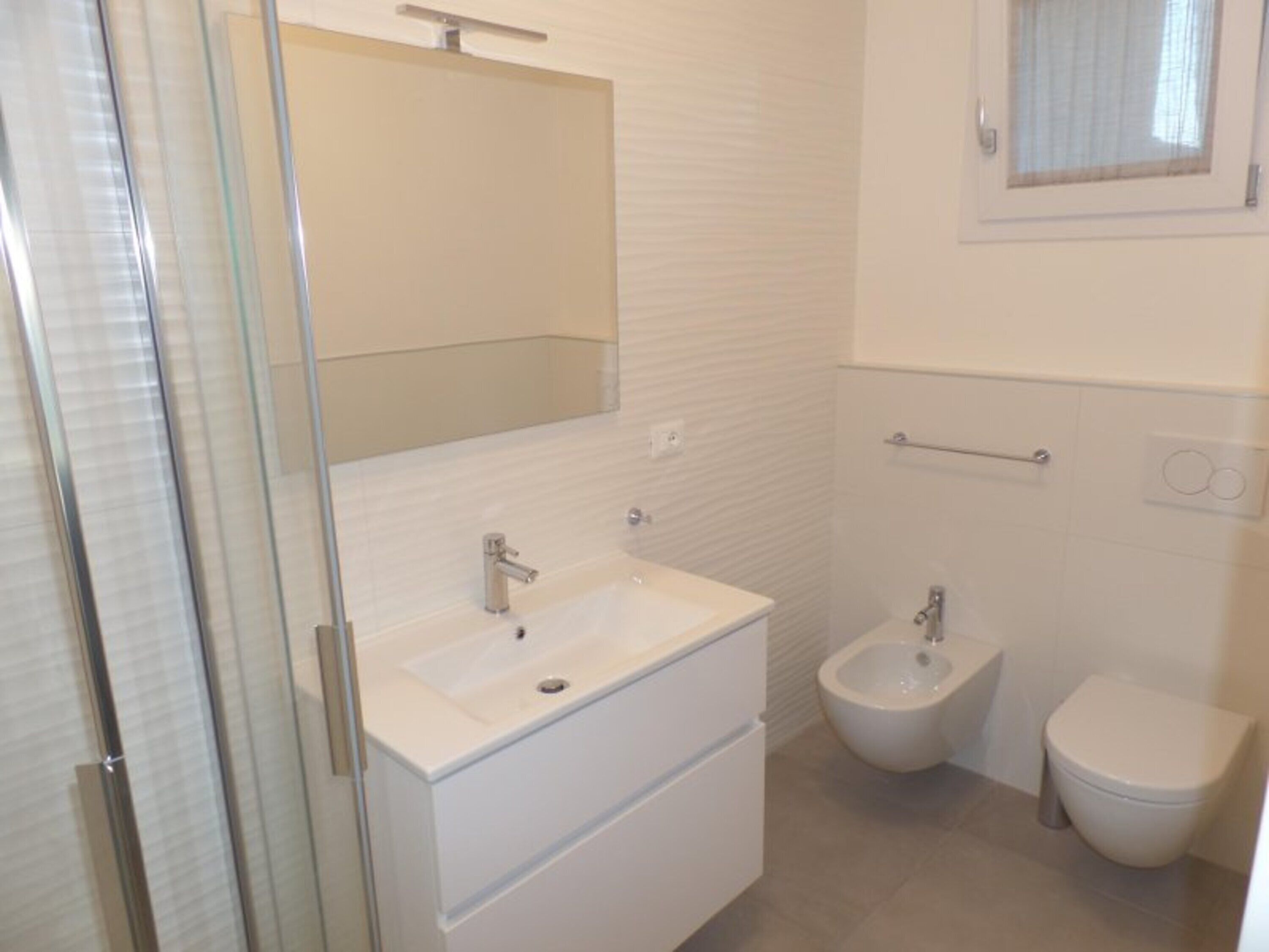 A bathroom with a glass shower enclosure, a white sink with a mirror above it, a bidet, and a toilet.