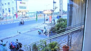 A view of a street with a building labeled 'NS GRAND' in the background, a 'BANQUET HALL LUXURY HALL LUXURY ROOMS' sign, and several potted plants on the sidewalk.