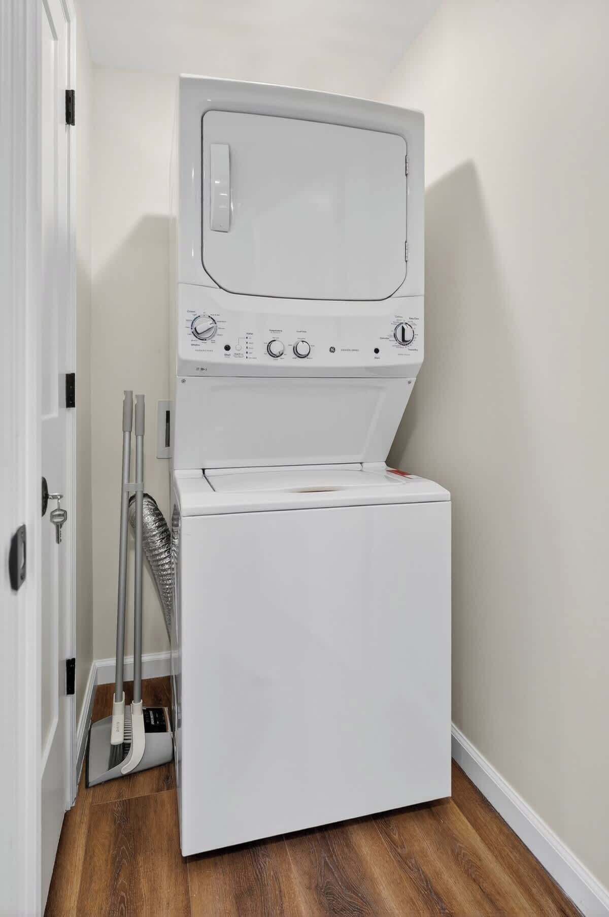 Une machine à laver et un sèche-linge blancs, placés dans un coin, avec un plancher en bois et une porte sur le côté gauche.