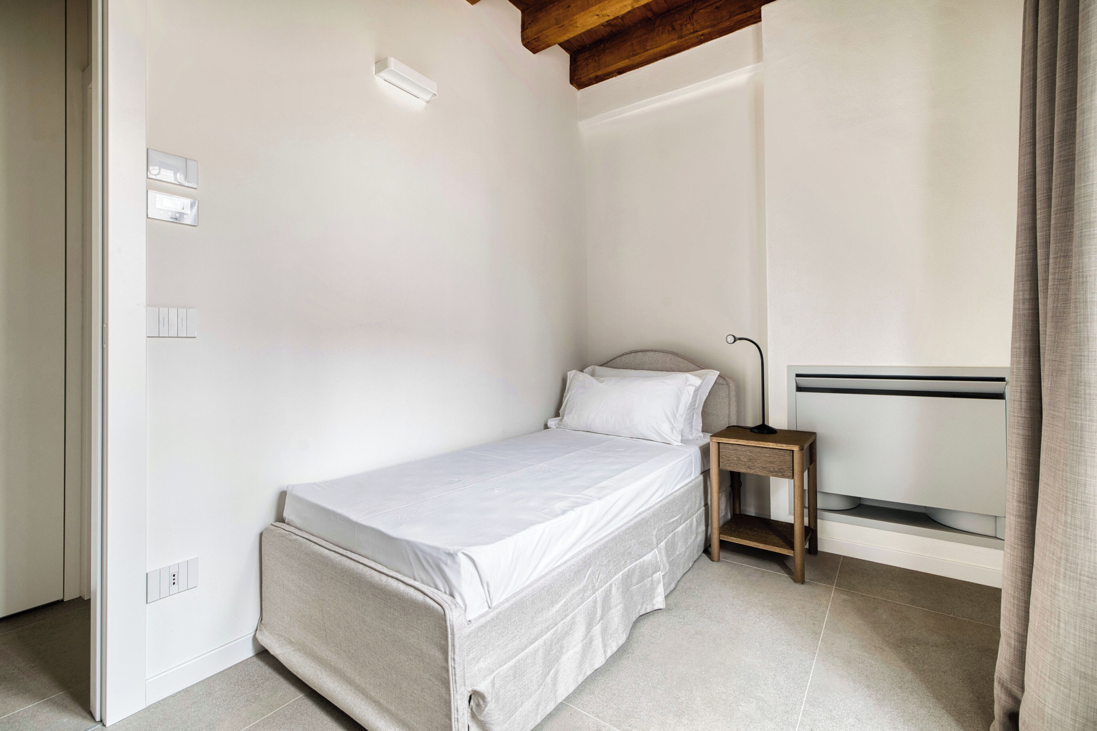 Ein kleines, sauberes Zimmer mit einem Bett, einem Nachttisch und einem Fenster mit Vorhängen.