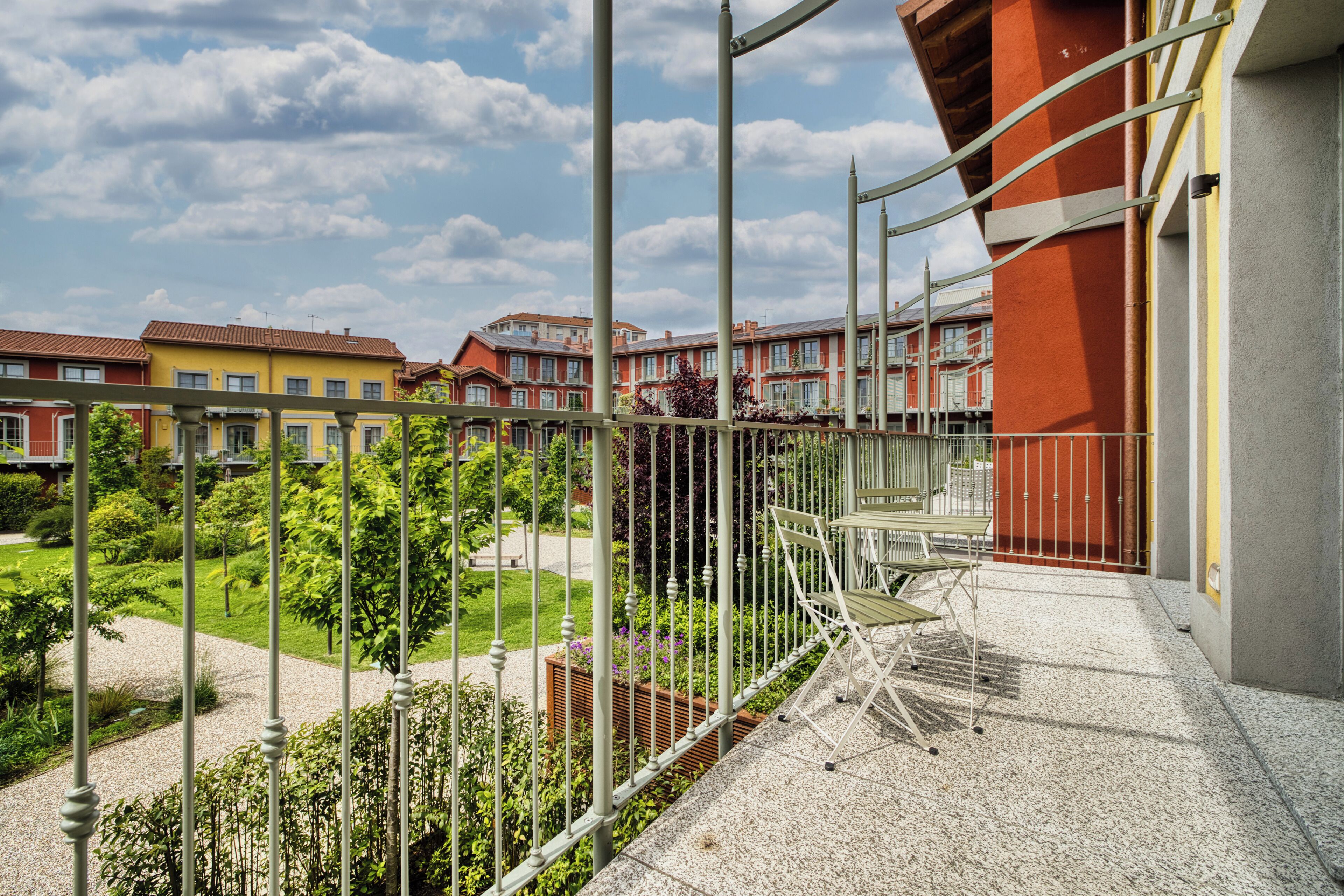 A balcony with a metal railing, a garden view, and a building with red and yellow facades.