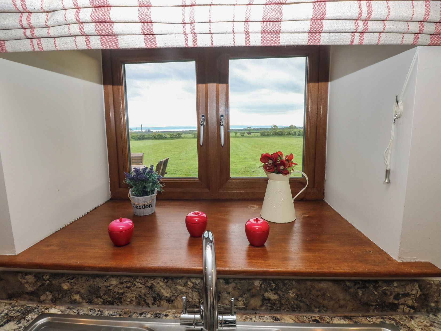 A kitchen with a window view of a green field and a white pitcher with red flowers on the counter.