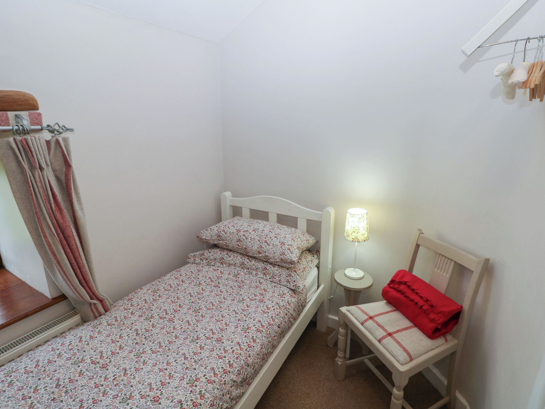A small, single-bed room with a white headboard, a bedside table with a lamp, a chair with a red blanket, and a curtain on the left side.