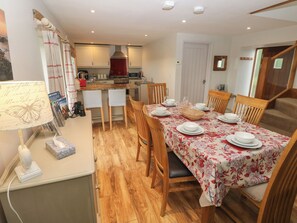 A dining area with a table set for four, wooden chairs, a kitchen in the background, and a wall-mounted lamp. - The Gallops (Barry)