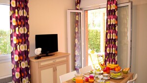 Une pièce avec un téléviseur posé sur un meuble en bois, une table à manger avec des aliments pour le petit-déjeuner, et des rideaux ornés d’un motif circulaire.