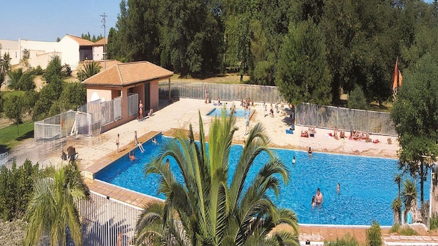 Une piscine entourée d’une zone délimitée par une clôture, où des personnes nagent et prennent des bains de soleil. On aperçoit des arbres en arrière-plan et un bâtiment avec un toit recouvert de tuiles.