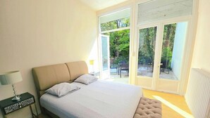Une chambre à coucher comprenant un lit, une table de chevet, une lampe et une porte-fenêtre coulissante donnant sur un espace extérieur aménagé avec des chaises.