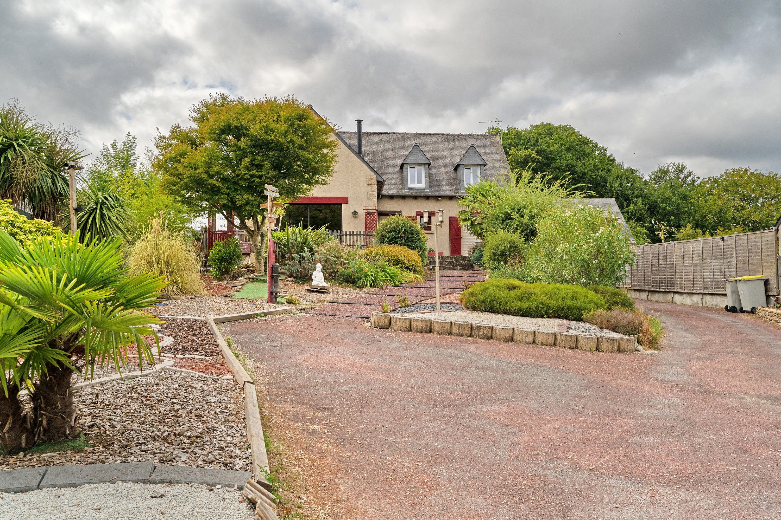 A house with a gravel driveway, surrounded by various plants and a statue.