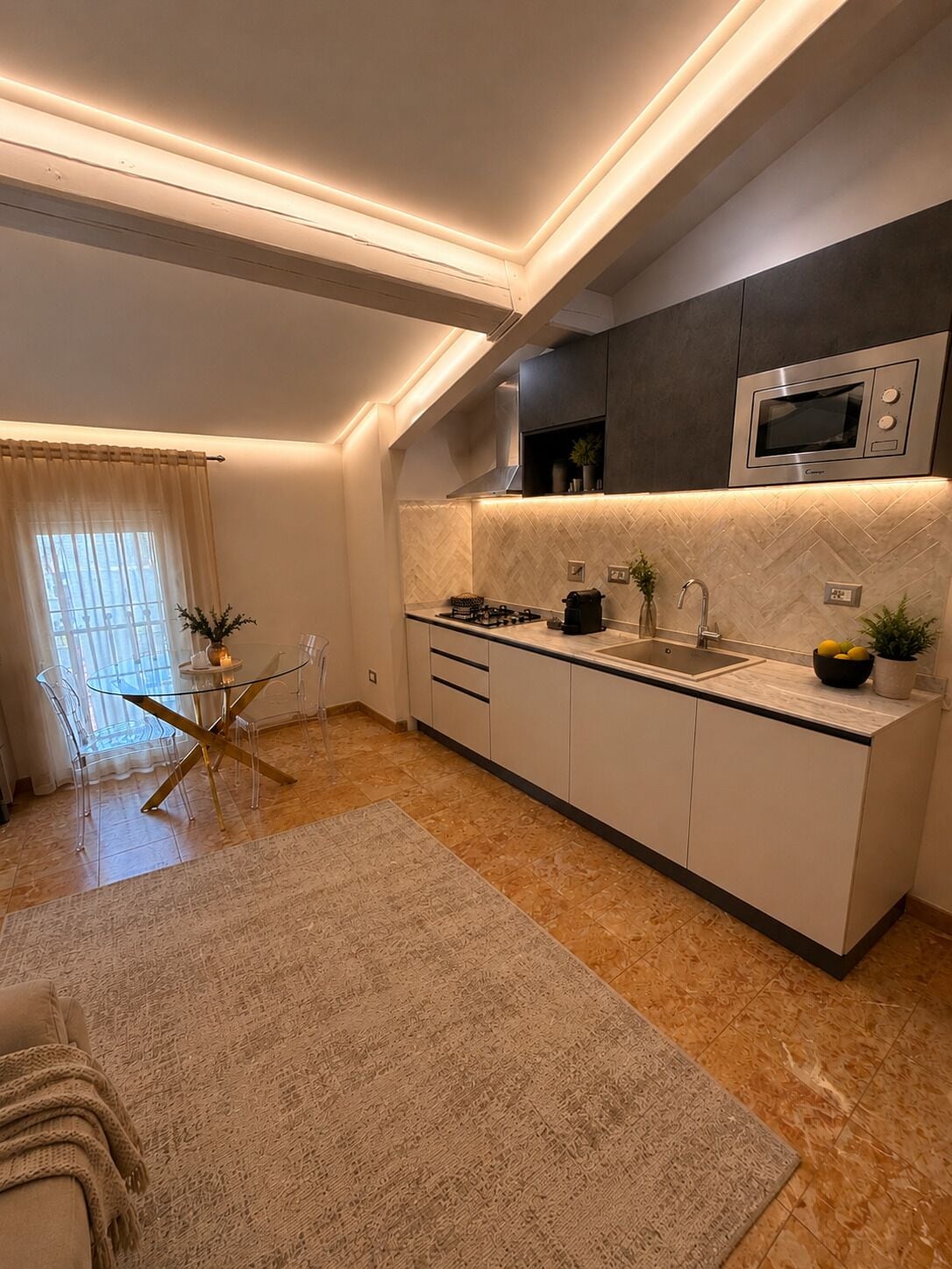 Een moderne keuken met een ingebouwde magnetron, een gasfornuis en een spoelbak. Er is een eethoek met een glazen tafel en stoelen, en een groot vloerkleed.