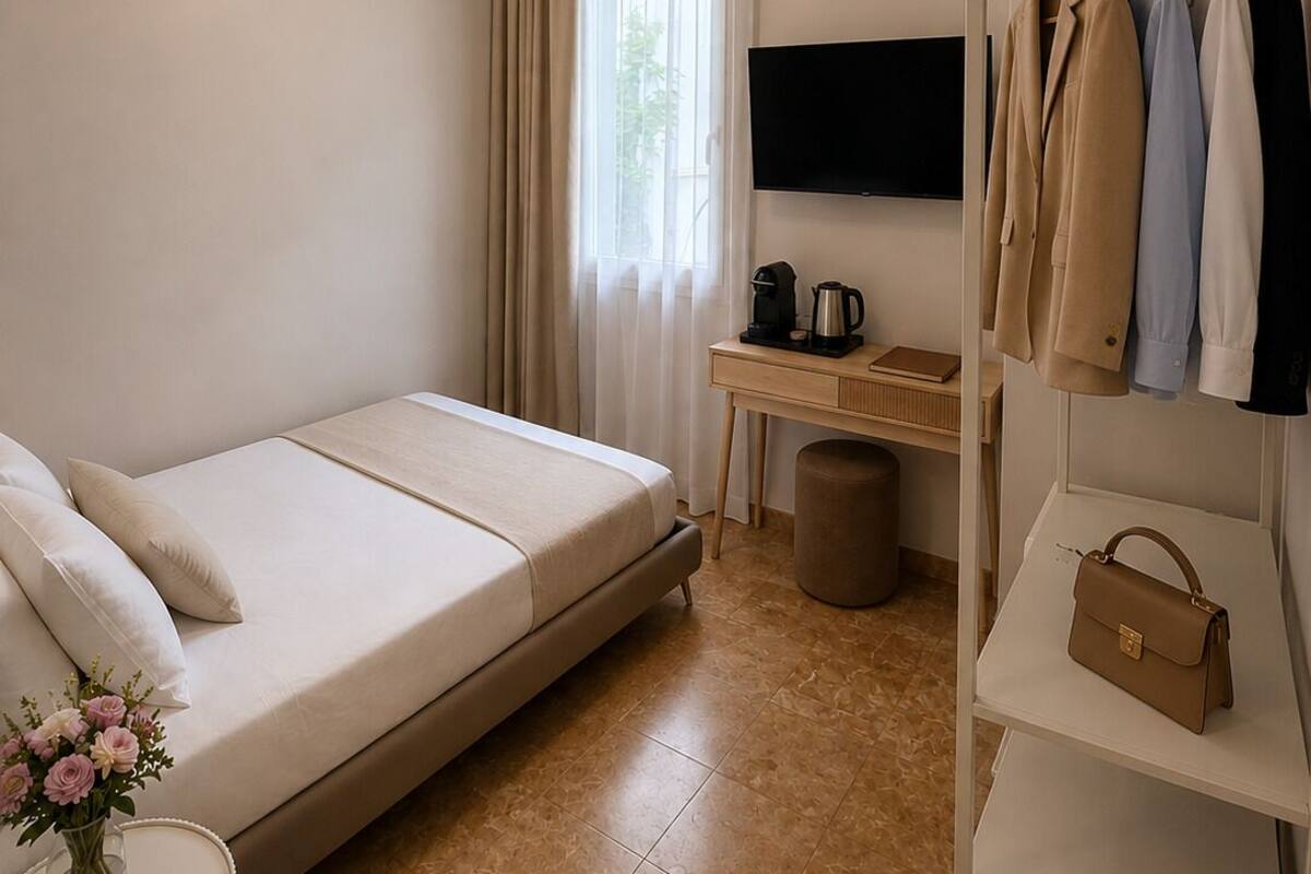 A hotel room with a bed, a desk with a flat-screen TV, a wardrobe, and a small table with a vase of flowers.