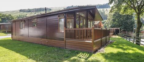 A wooden cabin with a deck, surrounded by grass and trees.