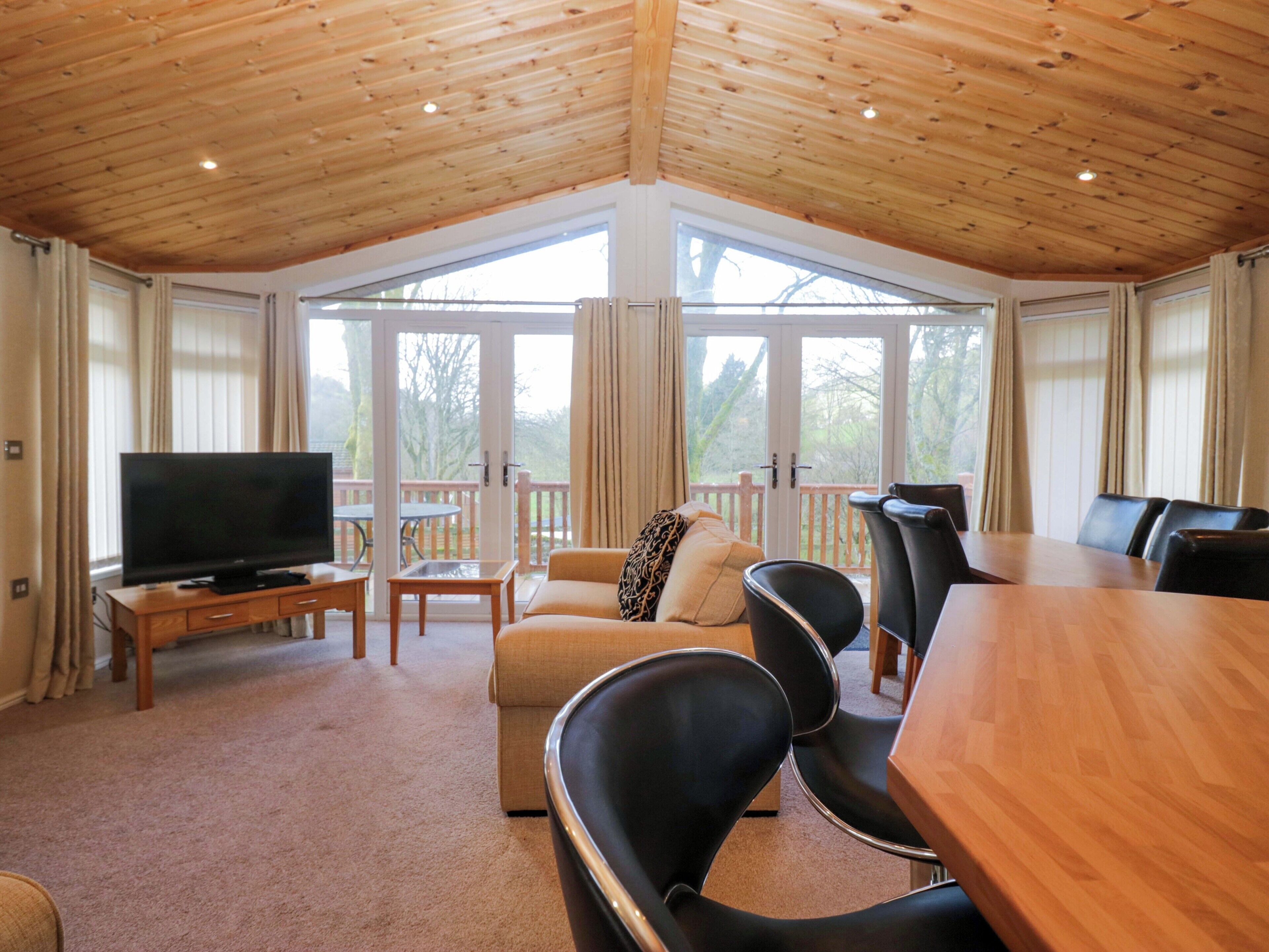 A room with a wooden ceiling, a television on a stand, a sofa, two black chairs, a wooden table, and large windows with curtains.