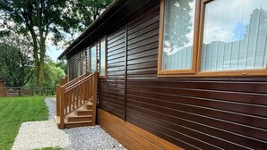 A wooden cabin with a wooden staircase leading to a door, surrounded by a gravel path and a grassy area. - Wally's Folly (Windermere)
