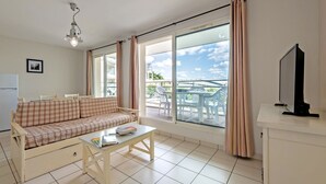 Una sala de estar con un sofá, una mesa de centro, un televisor y ventanas amplias con cortinas.