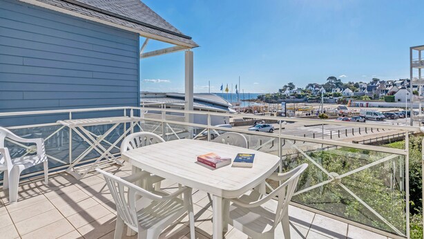 A balcony with a table and chairs, overlooking a coastal area with buildings and parked cars. - Balcon avec vue sur mer | Appt 6p + Accès  sauna (Plougonvelin)