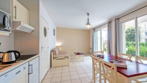 Een compacte keuken met een Bosch vaatwasser, een waterkoker en een kleine eettafel met stoelen. Er is een bed met een gestreept dekbed en een nachtkastje met een lamp. De kamer heeft een groot raam met uitzicht op groen.