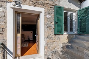 A stone building with a wooden door, a window with green shutters, and a staircase leading to the entrance.