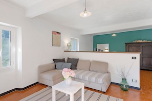 A living room with a sofa, a white table, a vase with flowers, a green wall, and a kitchen area in the background.