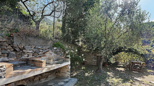 A stone-built outdoor kitchen with a grill, surrounded by trees and a stone wall.
