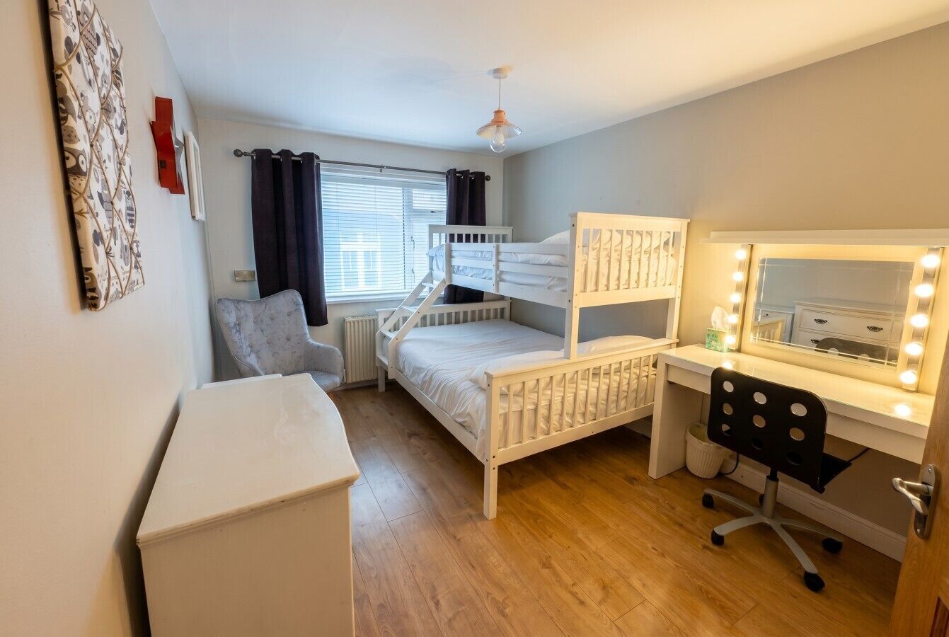 A bedroom with bunk beds, a desk with a mirror and lights, a chair, and a radiator.