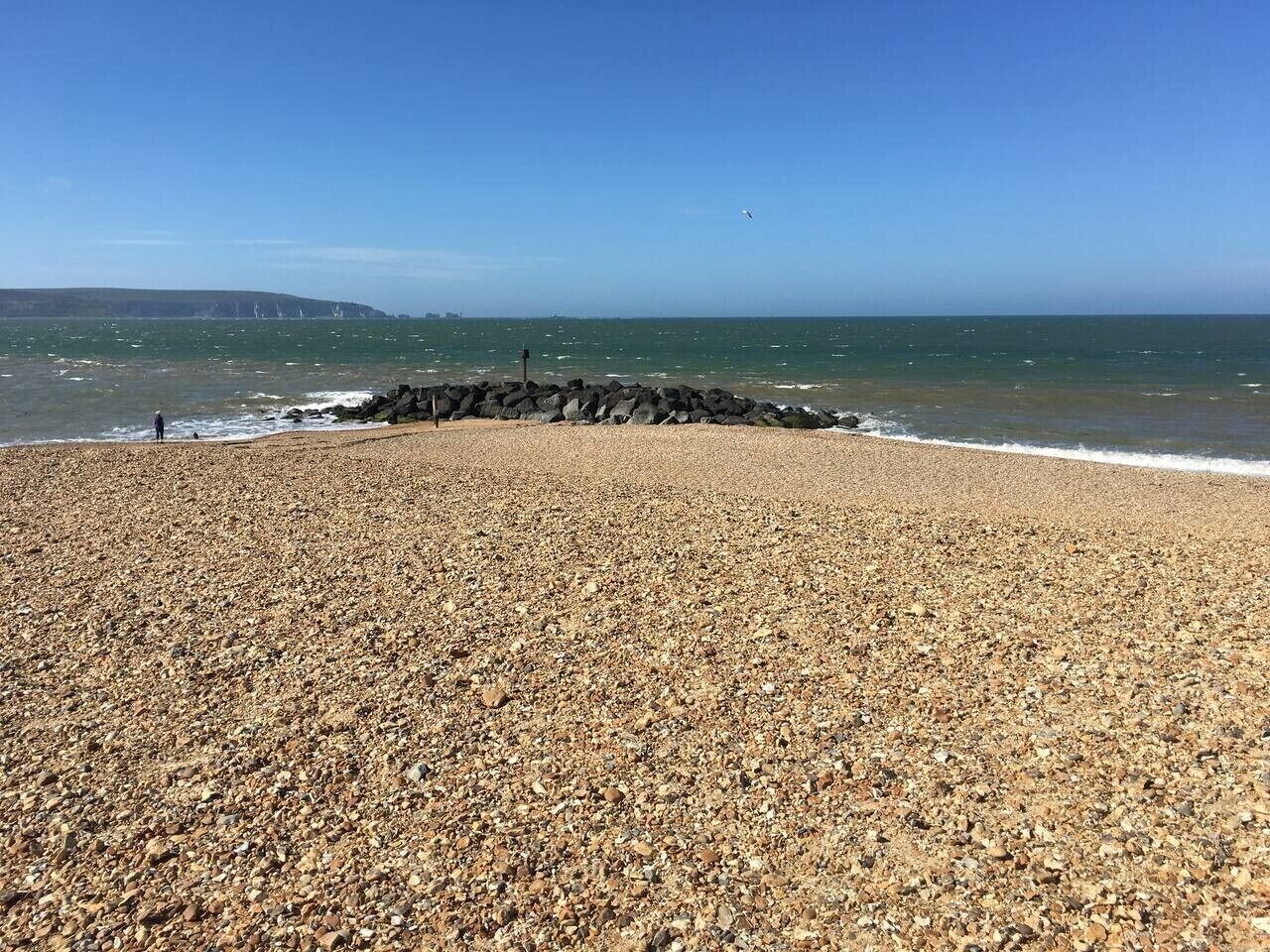 A pebbled beach with a rocky breakwater, clear blue sky, and calm sea.