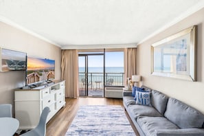 Ein Zimmer mit Fernseher, einer Couch, einem Esstisch und Meerblick durch große Fenster.