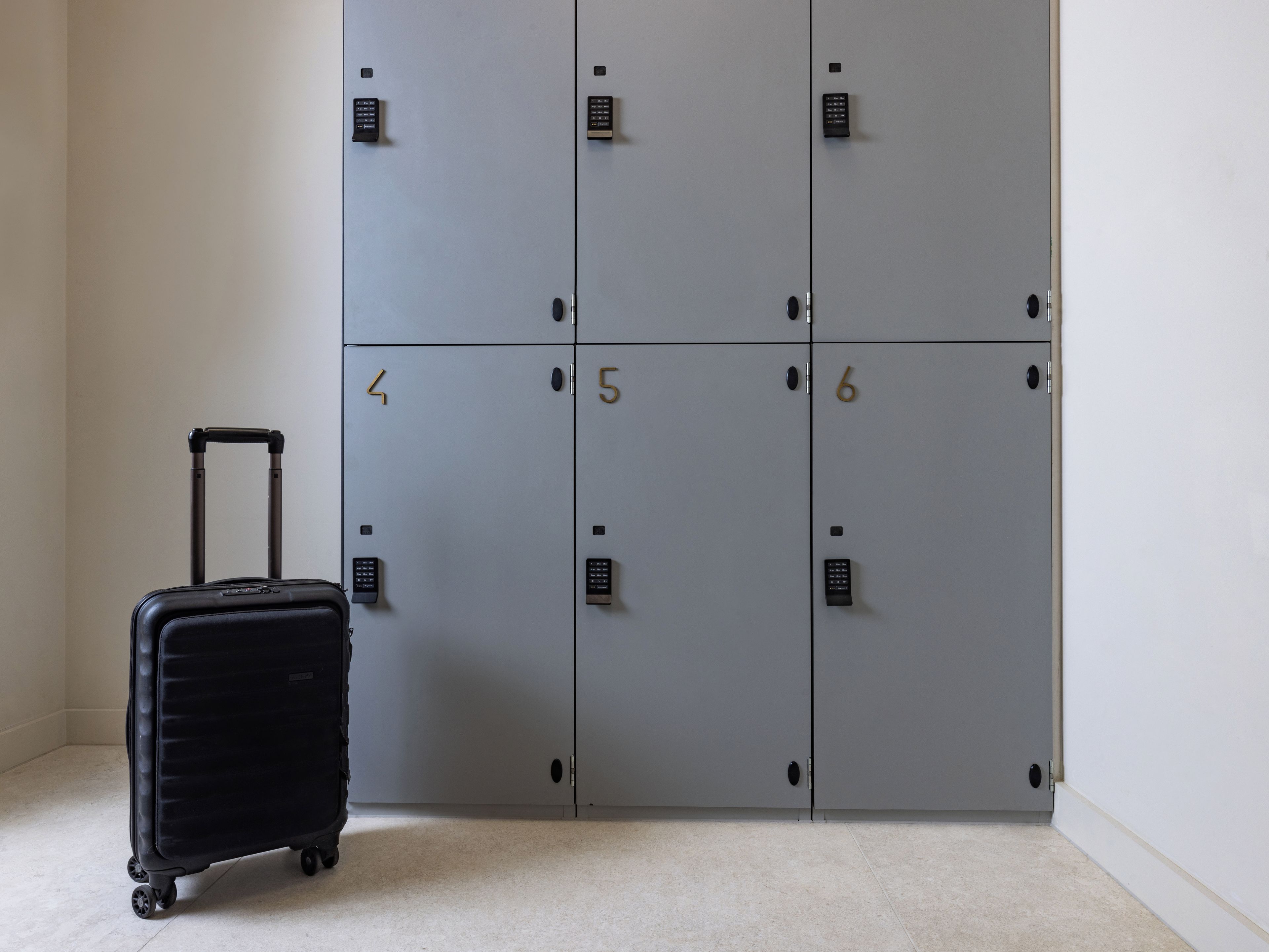 A row of grey lockers with numbers 4, 5, and 6, and a black suitcase with wheels in front of them.