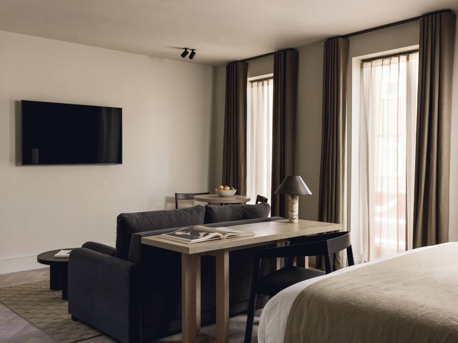 Quarto de hotel com cama, escrivaninha com luminária, sofá, televisão e cortinas.